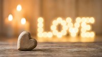 Wooden heart on rustic surface with glowing LOVE letters and candles in warm bokeh background, romantic Valentine's Day concept.