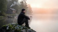 Young Caucasian man sitting on mossy rock by misty lake at sunrise, rustic cabin in foggy forest background, peaceful solitude.