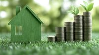 Green wooden house model with stacked coins and plant sprout on grass representing sustainable real estate investment and eco-friendly property growth concept.
