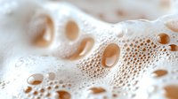 Creamy white foam bubbles in macro photography showing delicate texture and patterns of soap or beer froth with soft beige tones and circular shapes.