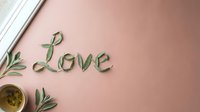 Word Love made from green leaves on a soft pink background with olive branch and tea cup, romantic flat lay for Valentine's Day.
