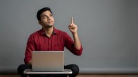Young South Asian male professional wearing burgundy button-up shirt, seated at desk with laptop, pointing upward with index finger, expressing idea or inspiration. Suitable for business, technology, education, and startup concepts.