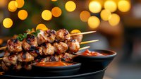 Grilled chicken skewers with sauce on black plate against blurred golden bokeh lights background, garnished with fresh herbs and served with dipping sauce.