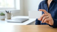Professional holding blank white business card at wooden desk with laptop and office supplies in bright workspace.