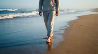 Woman walking barefoot on sandy beach at sunset with ocean waves and golden light reflecting on wet sand creating peaceful coastal atmosphere.