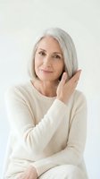 Mature woman with gray hair wearing cream knit sweater, hand to face in confident pose against white background for skincare and wellness marketing.