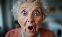 Senior Caucasian woman with surprised expression, wide blue eyes and open mouth showing genuine amazement. Close up portrait in orange sweater against blurred background.