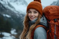 Young Caucasian woman hiker with long blonde hair wearing orange beanie and backpack smiling outdoors in snowy winter mountains during adventure trek.