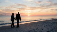 Two men walking together on beach at sunset holding hands in romantic moment with golden sky and ocean waves creating peaceful evening atmosphere.
