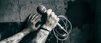Tattooed male hand gripping microphone with cable against textured concrete wall background for music performance and audio recording concepts.