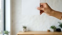 Close-up of hand with medium skin tone making thumbs down gesture against blurred office background with plants, expressing disapproval or negative feedback.