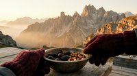 Hiker in burgundy gloves holds bowl of trail mix on wooden deck overlooking misty alpine peaks at sunrise, outdoor adventure and mountain trekking concept.
