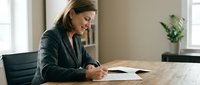 Professional businesswoman in dark suit signing documents at wooden desk in modern office environment for corporate and legal business concepts.