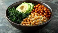 Healthy Buddha bowl with roasted chickpeas, fresh kale, sliced avocado and tahini dressing in rustic ceramic bowl on dark stone surface for nutritious meal prep.