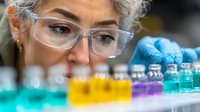 Senior female scientist in protective glasses examining colorful liquid samples in laboratory vials, conducting research and analysis in modern facility.