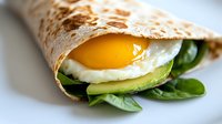 Fresh breakfast wrap with sunny side up egg, sliced avocado and baby spinach leaves on tortilla, close up view of morning meal on light background.
