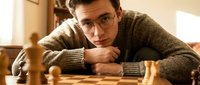 Young Caucasian man with glasses in brown sweater playing chess, thinking strategy in warm indoor setting with wooden pieces on board.