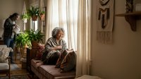 Senior woman reading book in cozy living room with natural light streaming through window while family member tends to plants in background.