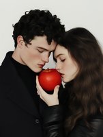 Young romantic couple in black clothing sharing intimate moment with red apple, symbolizing temptation and forbidden love against neutral background.