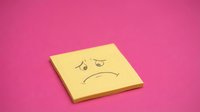 Yellow sticky note with sad face drawing and tear on bright pink background expressing disappointment and negative emotions for office communication.