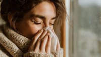 Young woman with cold or flu blowing nose into tissue while wearing cozy knit scarf near window with natural light.