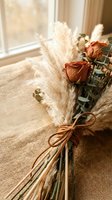 Dried flower bouquet with orange roses and pampas grass tied with leather cord on rustic burlap surface near window for home decor and wedding arrangements.