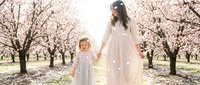 Woman and young girl holding hands in blooming orchard during spring season, wearing flowing white dresses, surrounded by flowering trees in natural daylight setting.