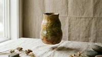 Handmade ceramic vase with organic green and brown glazed finish on linen tablecloth near window for home decor and artisan pottery display.