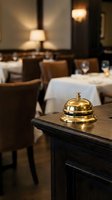 Golden service bell on wooden reception desk in upscale restaurant dining room with elegant table setting and warm ambient lighting for hospitality.