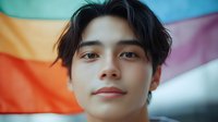 Young Asian man with dark hair smiling confidently against colorful rainbow pride flag background celebrating diversity and LGBTQ rights support.