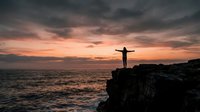 Silhouetted figure standing on rocky cliff with arms outstretched overlooking ocean at sunset, dramatic sky with orange and pink clouds, freedom and achievement concept for wellness and motivation.
