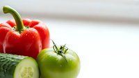 Fresh red bell pepper and green tomato with sliced cucumber on white surface for healthy cooking and meal preparation.