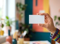 Young woman with medium skin tone holding blank white business card in creative studio workspace with colorful wall and plants, suitable for branding and corporate identity projects.