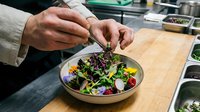 Hands of a male chef with medium skin tone garnishing a fresh salad bowl with microgreens and edible flowers in a professional kitchen setting for culinary content.