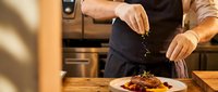 Professional chef in black apron seasoning grilled meat dish in modern restaurant kitchen. Culinary expertise and fine dining preparation concept.