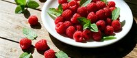 Fresh red raspberries with mint leaves arranged on white plate and wooden surface. Ideal for food, nutrition, healthy eating, and culinary content.