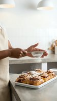 Person with dark skin tone dusting powdered sugar onto freshly baked cinnamon rolls in modern kitchen. Baking and pastry preparation concept for culinary content.