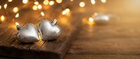 Two silver heart-shaped Christmas ornaments on rustic wooden surface with warm golden bokeh lights creating festive holiday atmosphere for seasonal decorations.