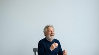 Senior man with gray curly hair and beard, wearing dark navy sweater, gesturing while speaking with animated expression against white background for communication and presentation concepts.
