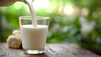 Fresh milk pouring into glass on rustic wooden table with green nature background. Healthy dairy product for breakfast and nutrition concepts.