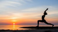 Woman practicing yoga warrior pose silhouette at sunrise by ocean beach. Peaceful morning meditation and fitness routine with golden sky reflection on water.