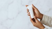 Hands of a woman with medium skin tone holding a white cosmetic bottle against a marble background, suitable for skincare, beauty product marketing, and wellness advertising.