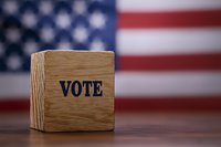 Wooden cube with VOTE text against blurred American flag background on dark wooden table, symbolizing democracy and electoral process in USA.