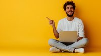 Young Middle Eastern man with curly hair sitting cross-legged on floor with laptop, wearing casual white outfit, pointing sideways against vibrant yellow background.