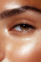 Close-up of brown eye with glowing glitter makeup, long eyelashes, and defined eyebrow on mixed race female model with freckles and dewy skin texture.