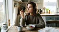Young woman enjoying healthy breakfast bowl in bright modern kitchen, sitting at wooden table with coffee cup and newspaper, morning lifestyle concept.