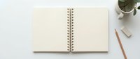 Open blank spiral notebook with cream pages on white desk surrounded by pencil, eraser and potted plant for writing planning and creative work.