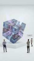 Transparent geometric cube sculpture with purple and blue crystalline blocks stacked in abstract formation, displayed on white platform with visitors viewing contemporary art installation.