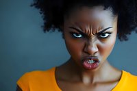 African American woman with angry facial expression showing intense emotions and furrowed brow wearing bright yellow top against blue background.