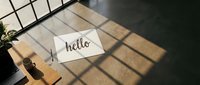 White card with hello lettering on wooden desk in sunlit modern loft interior with window shadows and concrete floor.